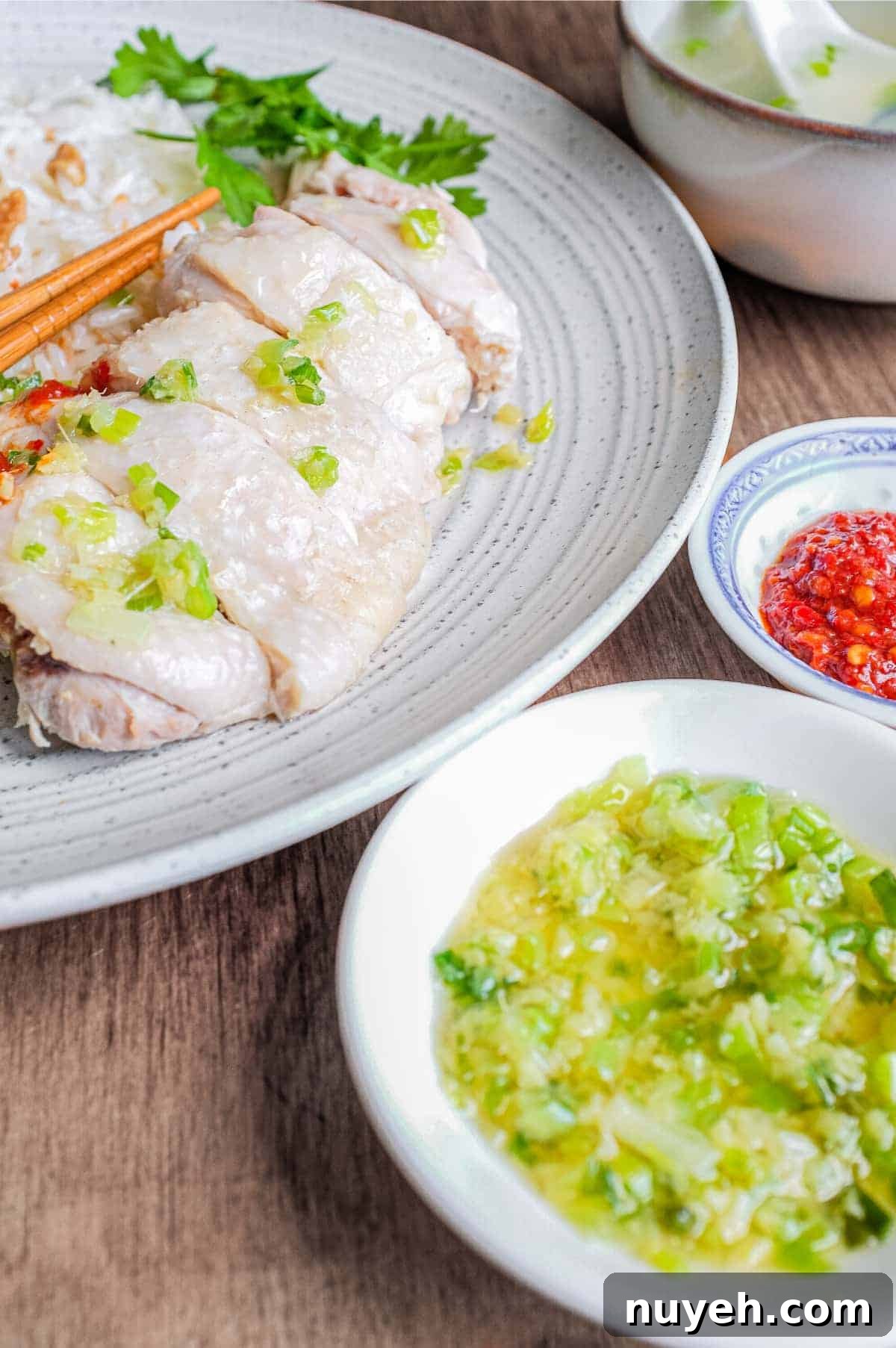 A plate of Hainan Chicken Rice with chopsticks. Featuring poached chicken, rice with specks of chicken skin, chopped cucumber and a sprig of cilantro. A small bowl of soup and an asian spoon sits to the side along with two condiments of chilli sauce and a ginger scallion sauce.