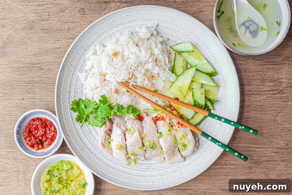 A plate of Hainan Chicken Rice with chopsticks. Featuring poached chicken, rice with specks of chicken skin, chopped cucumber and a sprig of cilantro. A small bowl of soup and an asian spoon sits to the side along with two condiments of chilli sauce and a ginger scallion sauce.