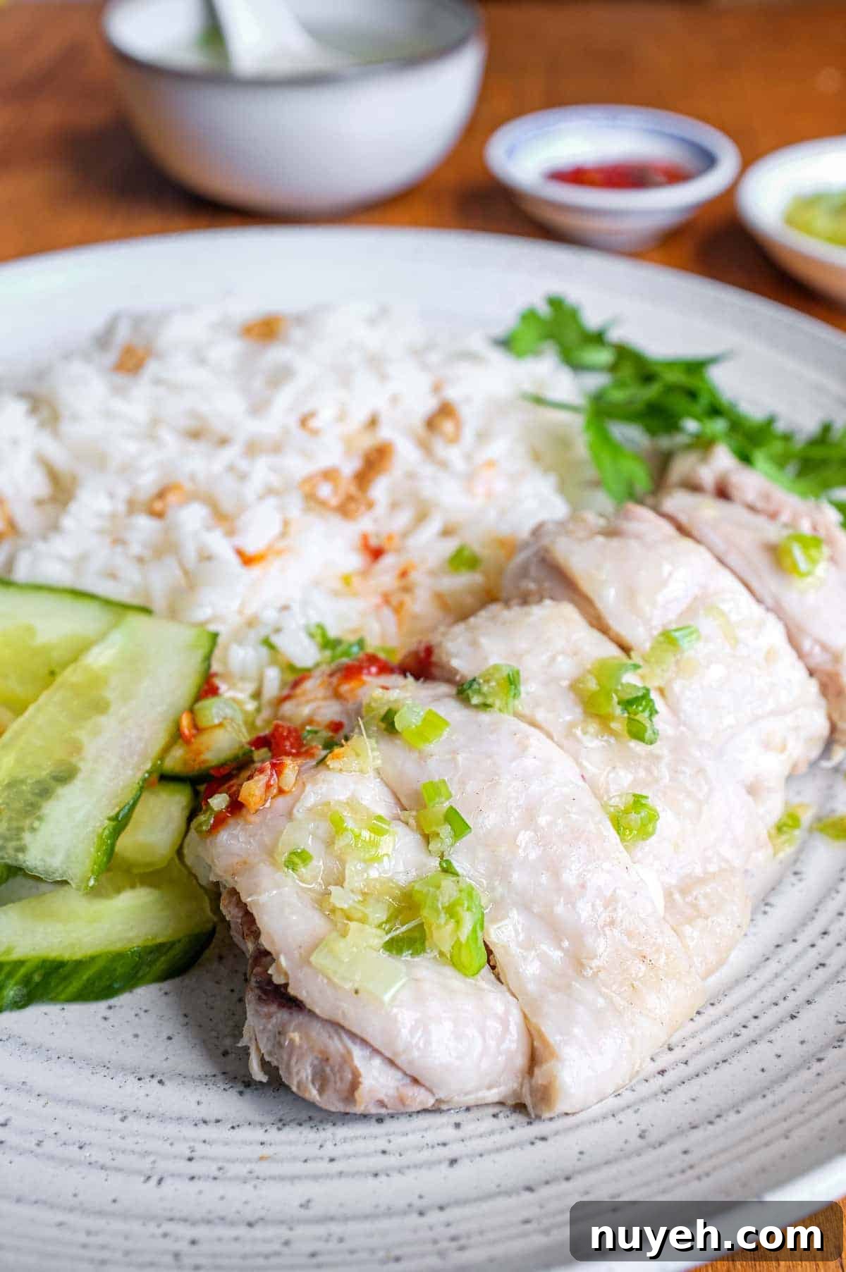A plate of Hainan Chicken Rice with chopsticks. Featuring poached chicken, rice with specks of chicken skin, chopped cucumber and a sprig of cilantro. A small bowl of soup and an asian spoon sits to the side along with two condiments of chilli sauce and a ginger scallion sauce.