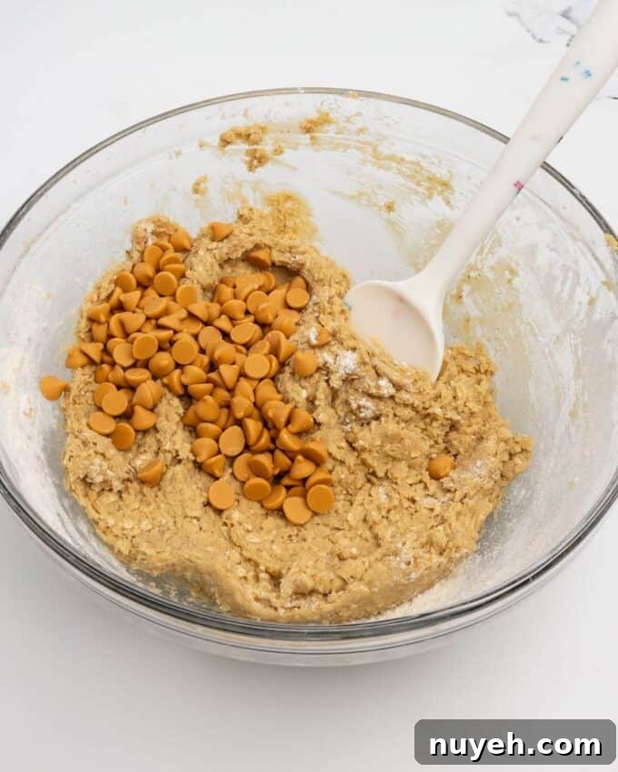 adding butterscotch chips to oatmeal scotchie dough in glass mixing bowl with rubber spatula