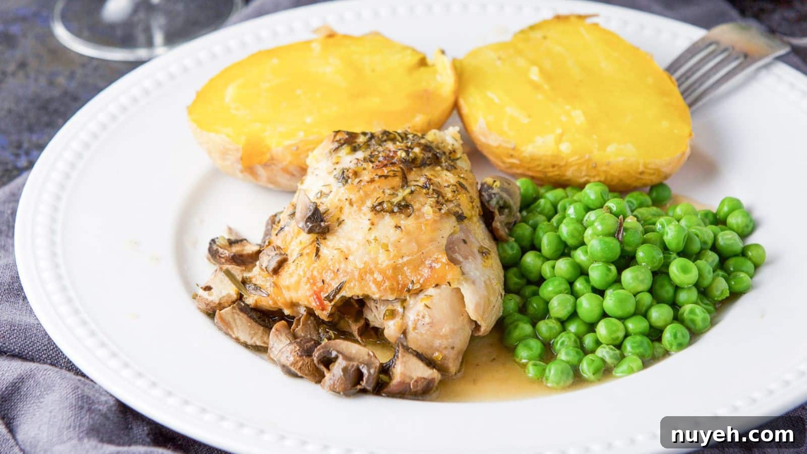 A white beaded plate with a chicken thigh with mushrooms and sauce, peas and baked potato