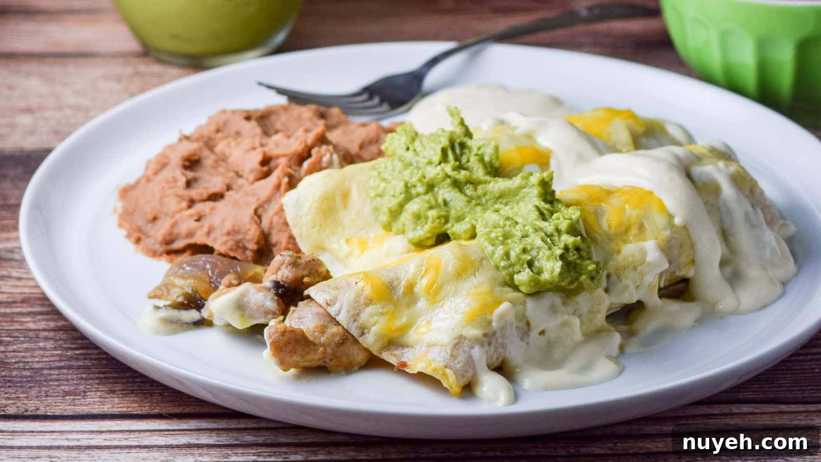A white plate with two enchiladas with chicken sticking out of it with sauce and guacamole - along with beans on the plate