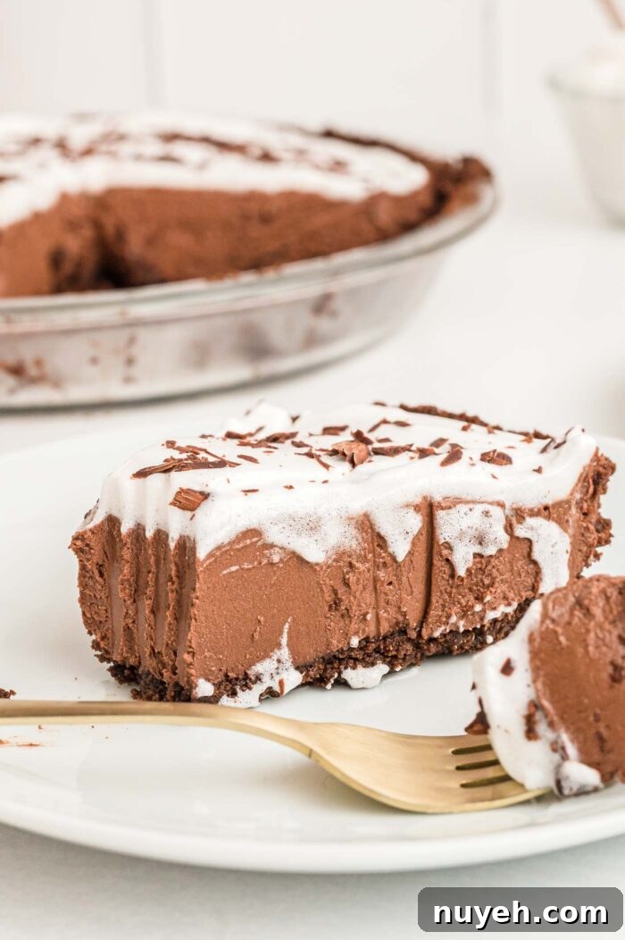 A slice of chocolate pie with chocolate crust topped with whipped cream and chocolate shavings on a small plate. The slice has a bite taken from it and you can see the marks a fork made when it sliced through the pie. A fork with some pie on it rests on the plate.