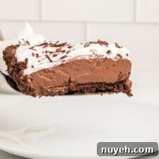 A slice of vegan chocolate pie with a chocolate crust topped with whipped cream on a spatula held over a plate against a white subway tile background.