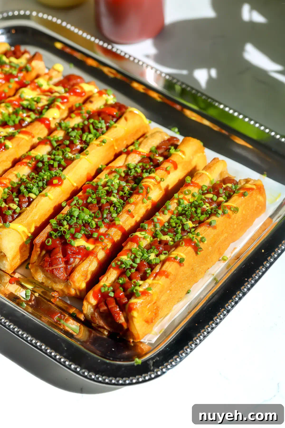 Crescent roll hot dogs on a silver tray topped with spicy ketchup, mustard mayo, and chopped chives.