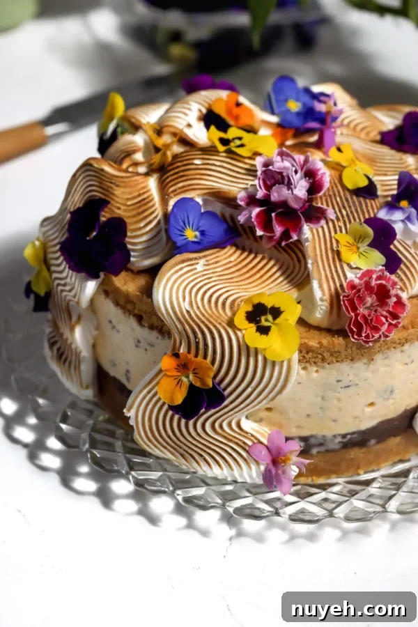 S'mores ice cream cake on a glass cake stand topped with torched Italian meringue in waves and edible flowers, with the chocolate ganache layer and graham crust visible.