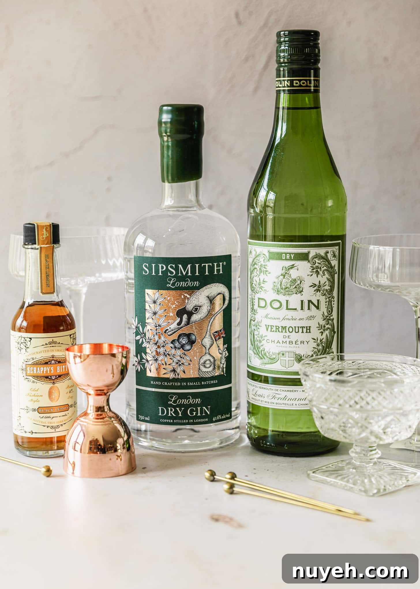 A bottle of gin, bottle of vermouth, bottle of lemon bitters, glasses, and copper shot glass on a pink marble counter.