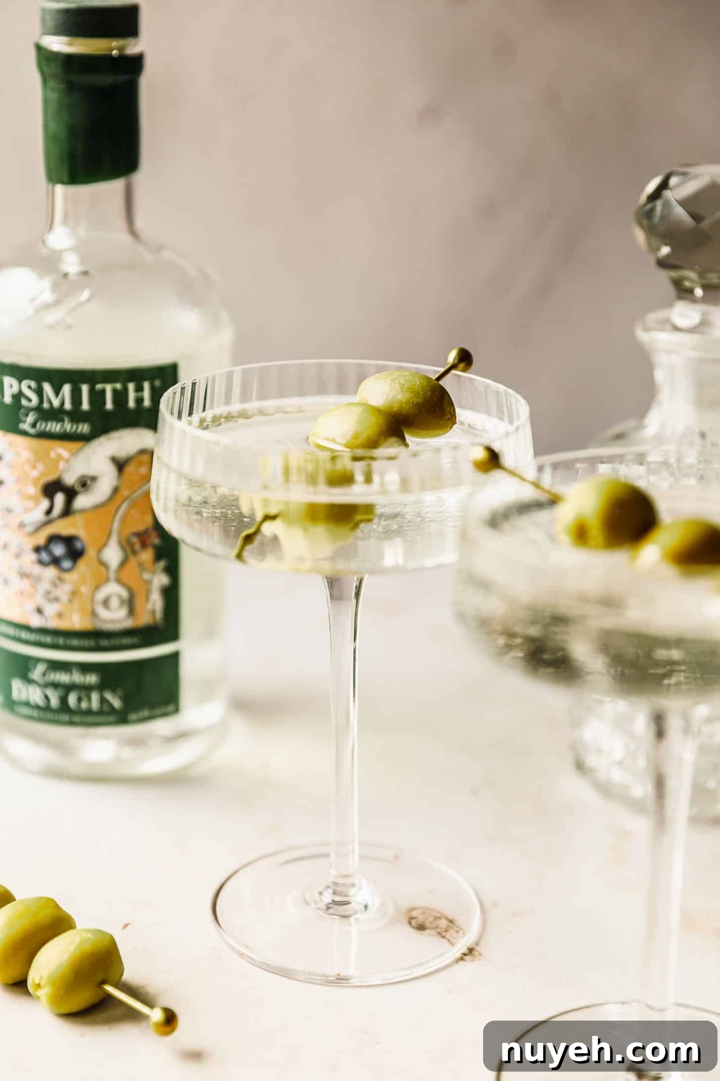 Two freezer door martinis on a tan marble counter next to a bottle of gin, decanter, and gold pick with olives.