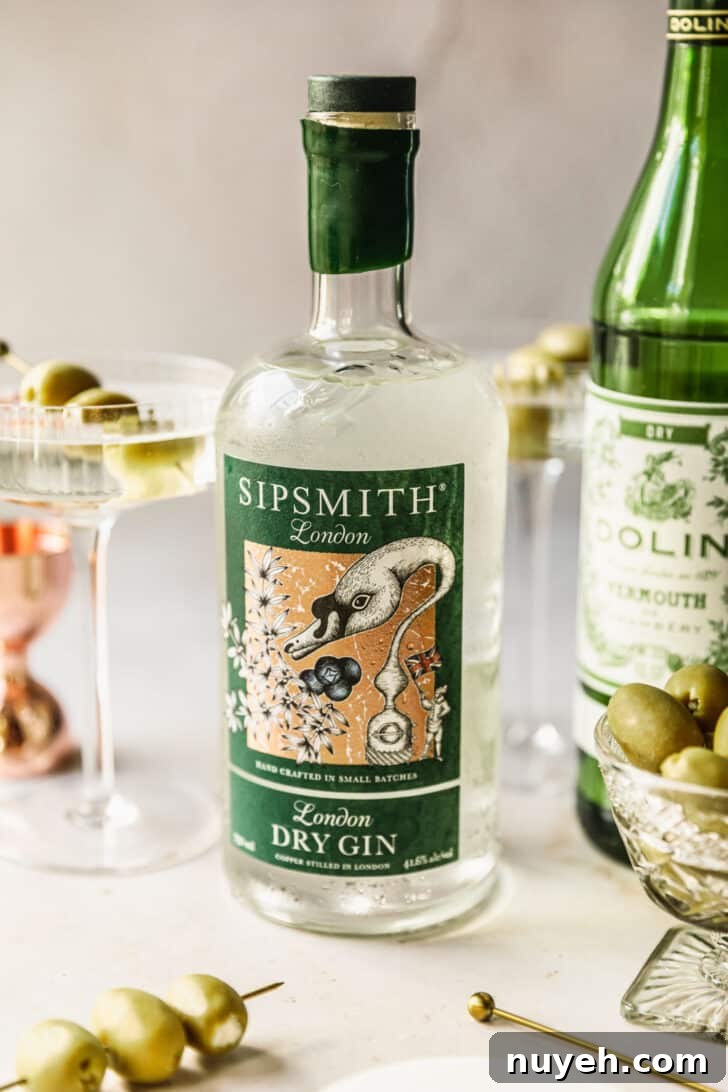 A bottle of gin on a tan marble counter next to a bottle of vermouth, cocktail glass, copper shot glass, and glass bowl of olives.