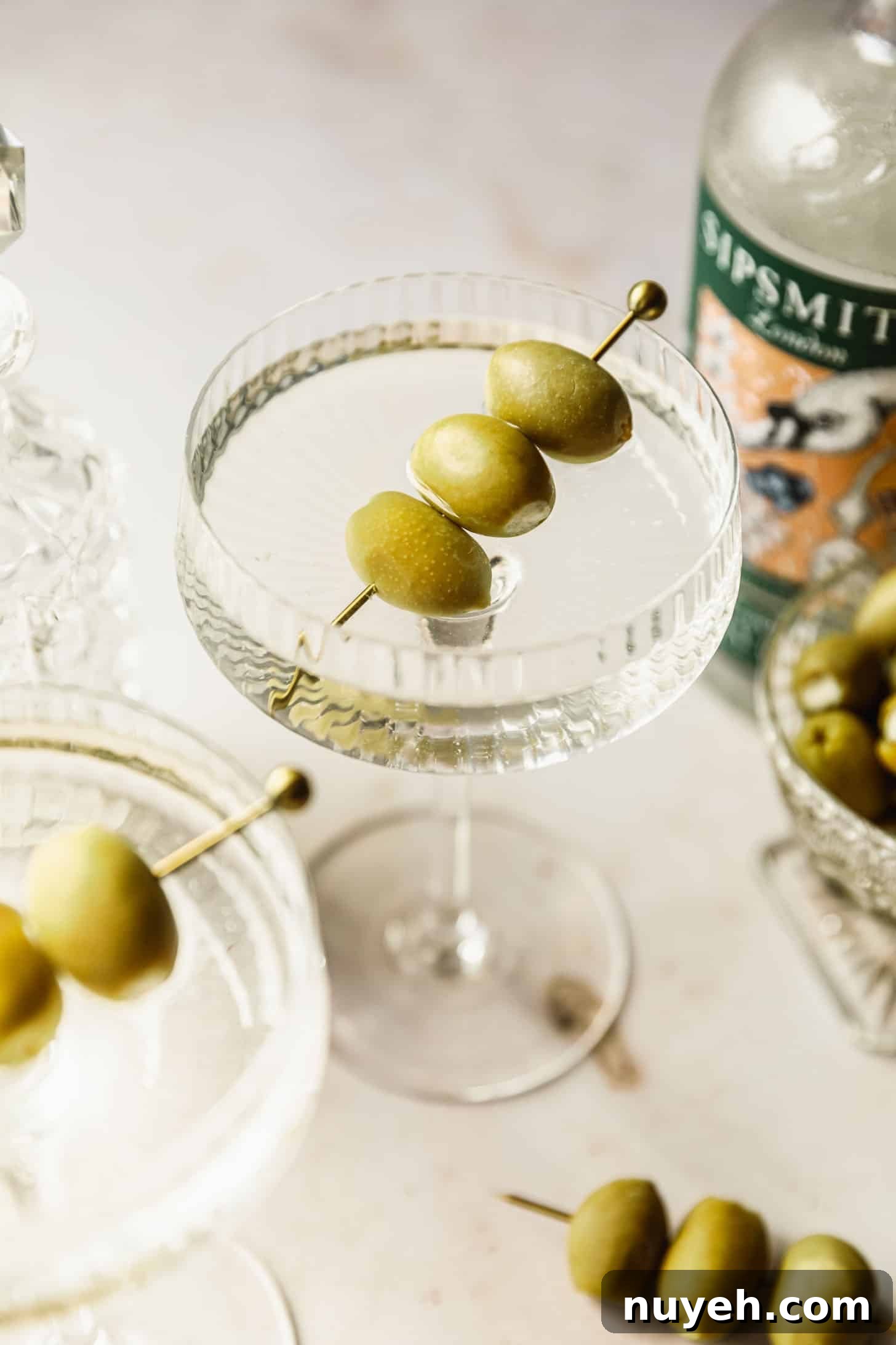 Two freezer door martinis with olives on a tan marble counter next to a glass bowl of olives, bottle of gin, and gold pick of olives.