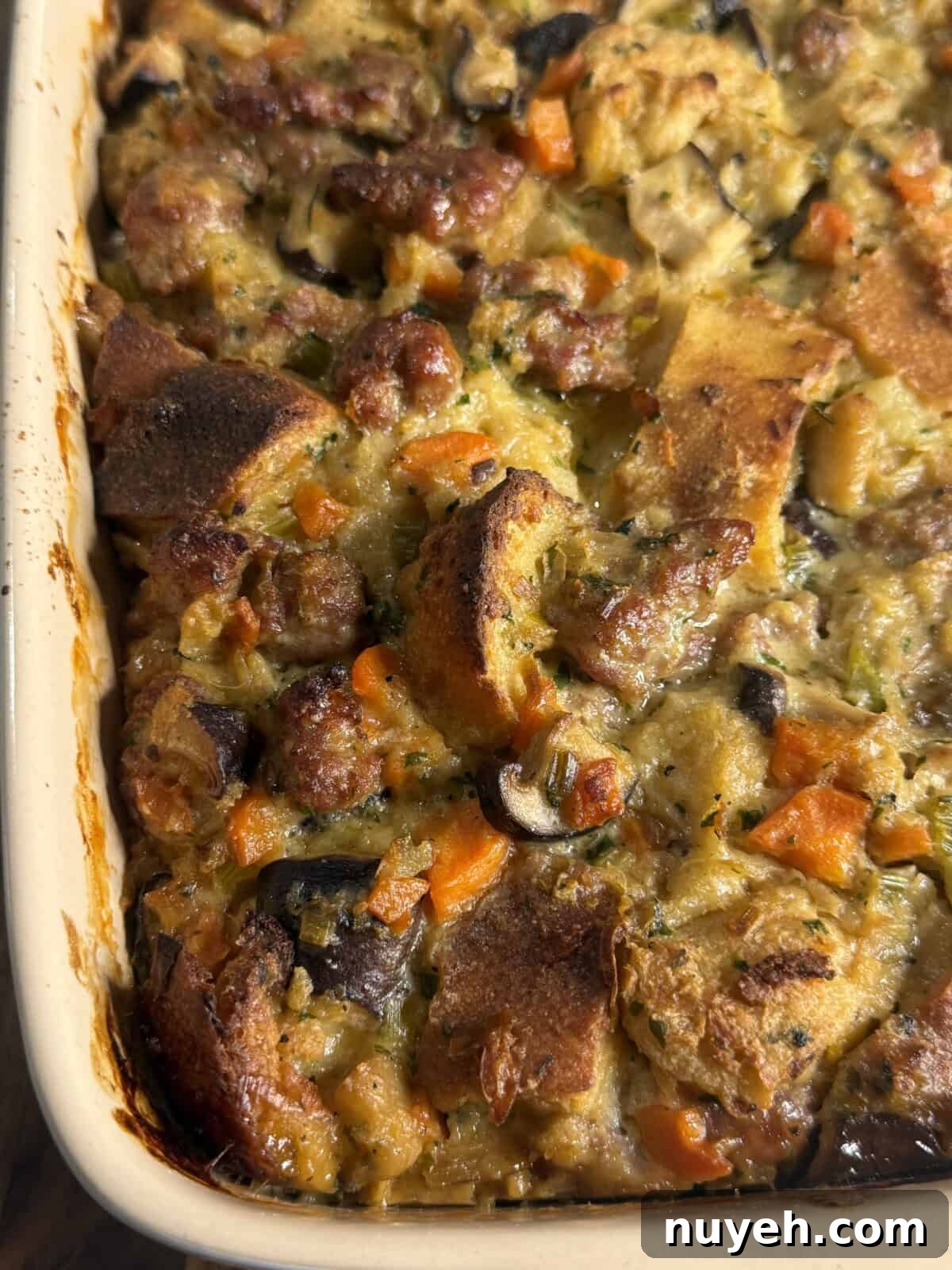 Sausage and Leek Bread-Pudding-Style Stuffing 2 A close-up of a baked casserole in a rectangular dish, featuring chunks of bread, sausage, carrots, mushrooms, and herbs, with a golden-brown, crispy top.