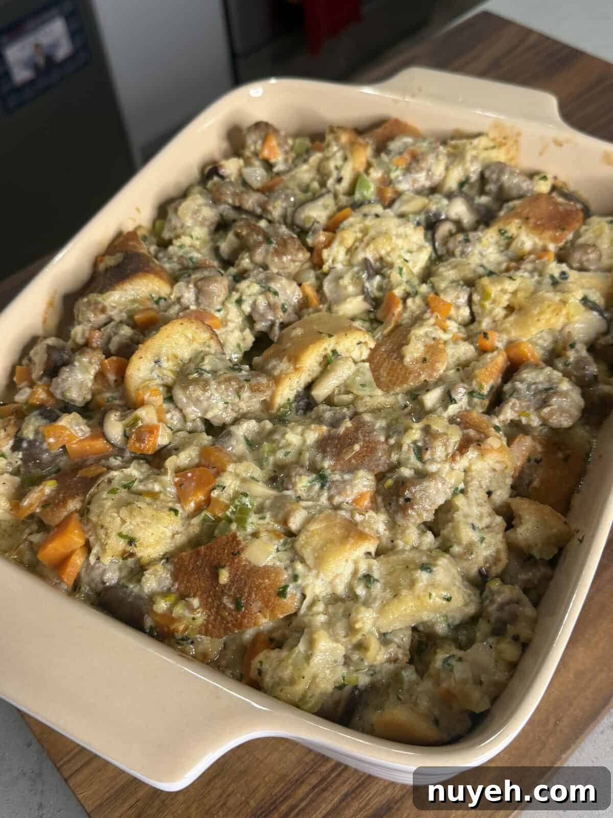 Sausage and Leek Bread-Pudding-Style Stuffing 3 A casserole dish filled with homemade stuffing, featuring chunks of bread, sausage, carrots, mushrooms, and herbs, sitting on a wooden surface in a sunlit kitchen.