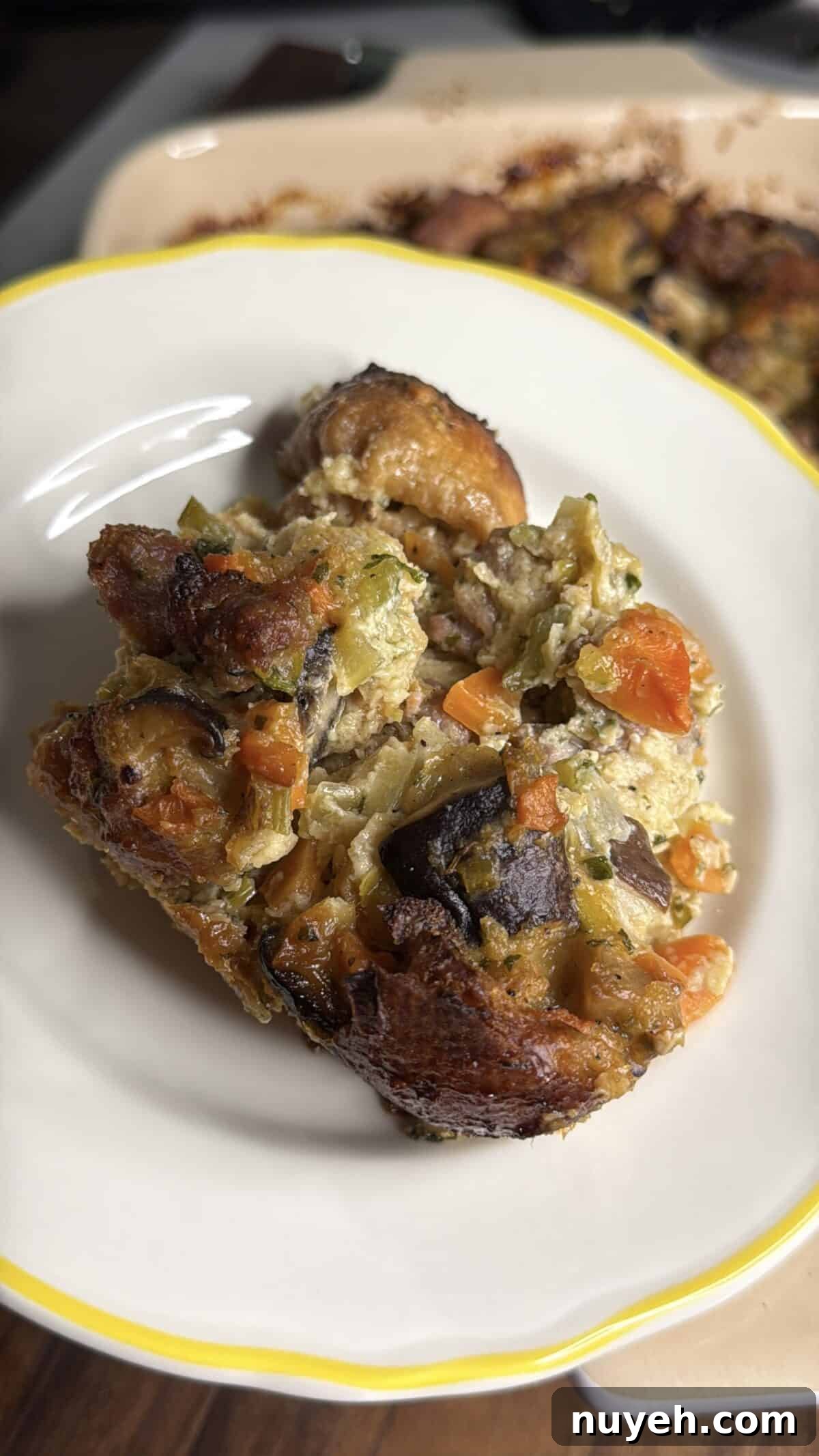 Sausage and Leek Bread-Pudding-Style Stuffing 4 A close-up of a serving of vegetable bread pudding, featuring chunks of bread, carrots, celery, eggplant, and other mixed vegetables on a white plate with a yellow rim.