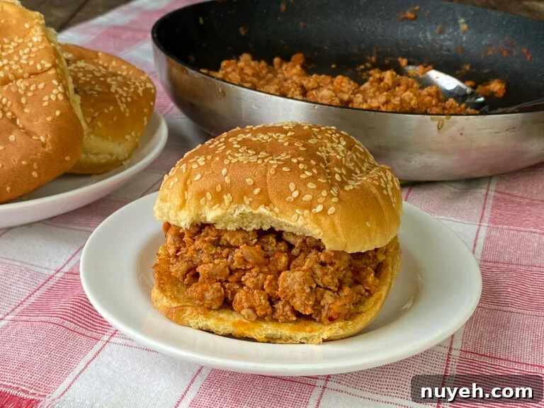 Sloppy Jane recipe in a sandwich on a white plate and red plaid napkin next to skillet of meat and extra buns