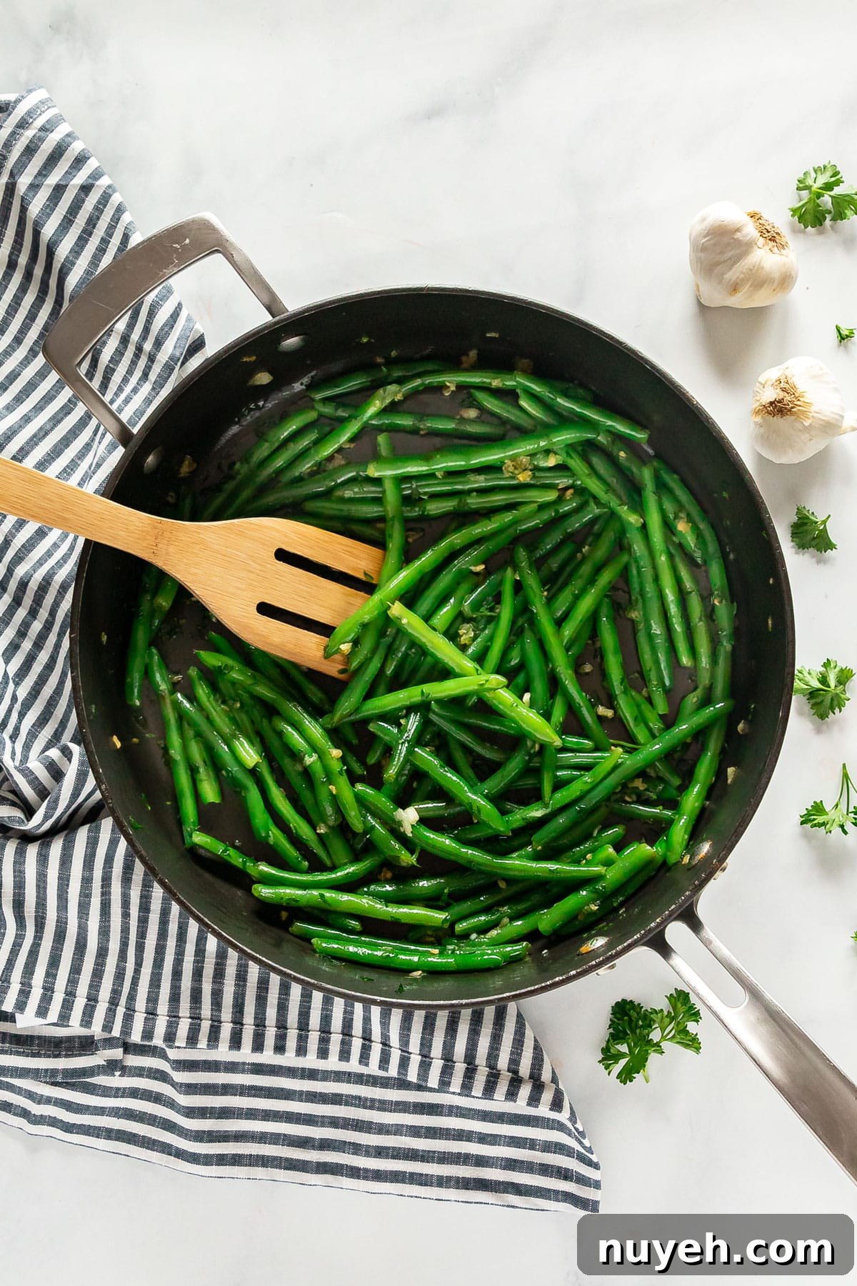 An overhead image of a pan of garlic green beans with a wooden spatula in the pan and with a stripped towel, garlic and parsley around it.