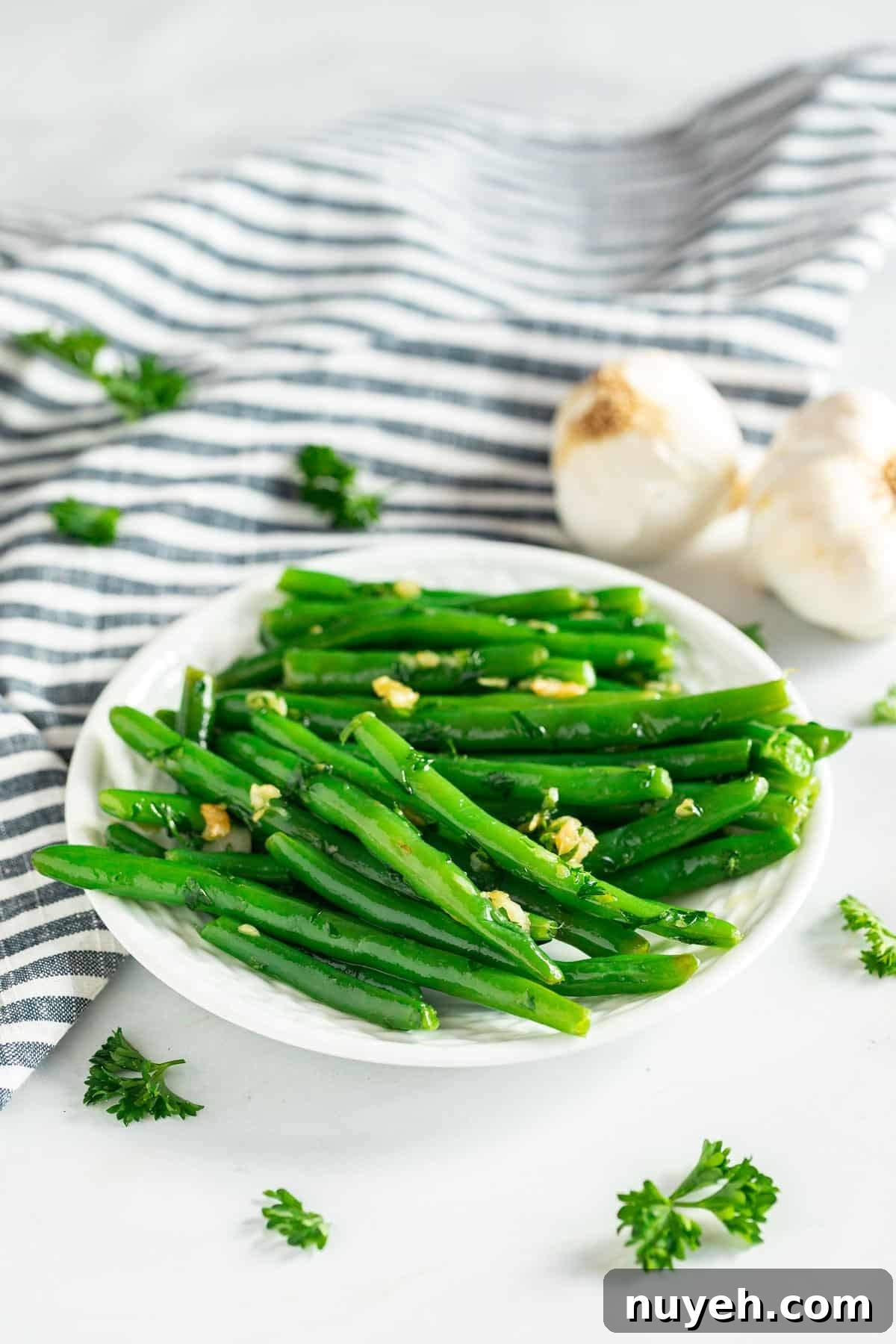 A plate of garlic green beans  with a striped towel, garlic bulbs and parsley in the background.