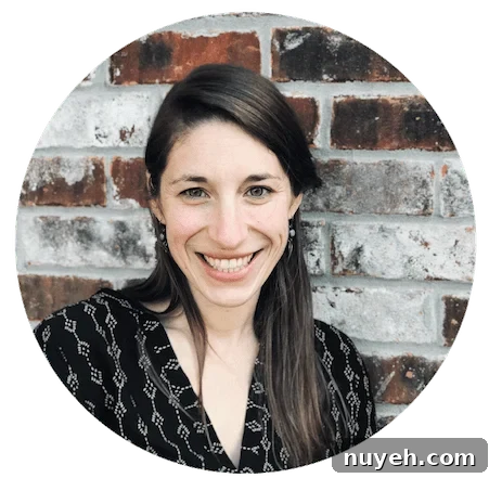 A brunette with straight hair and green earrings wearing a gray shirt in front of a red and white brick wall.