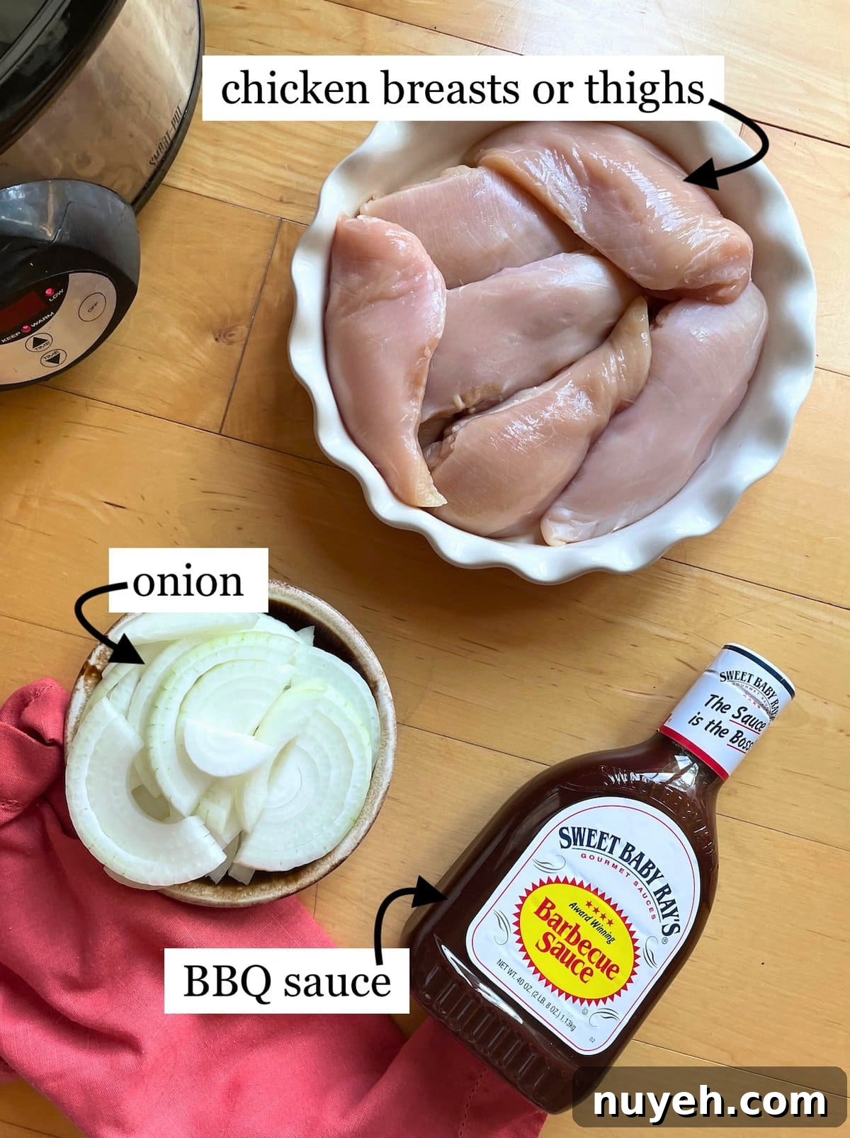 A bowl of raw chicken breasts or thighs, sliced onions, and a bottle of BBQ sauce on a wooden table alongside a crockpot.