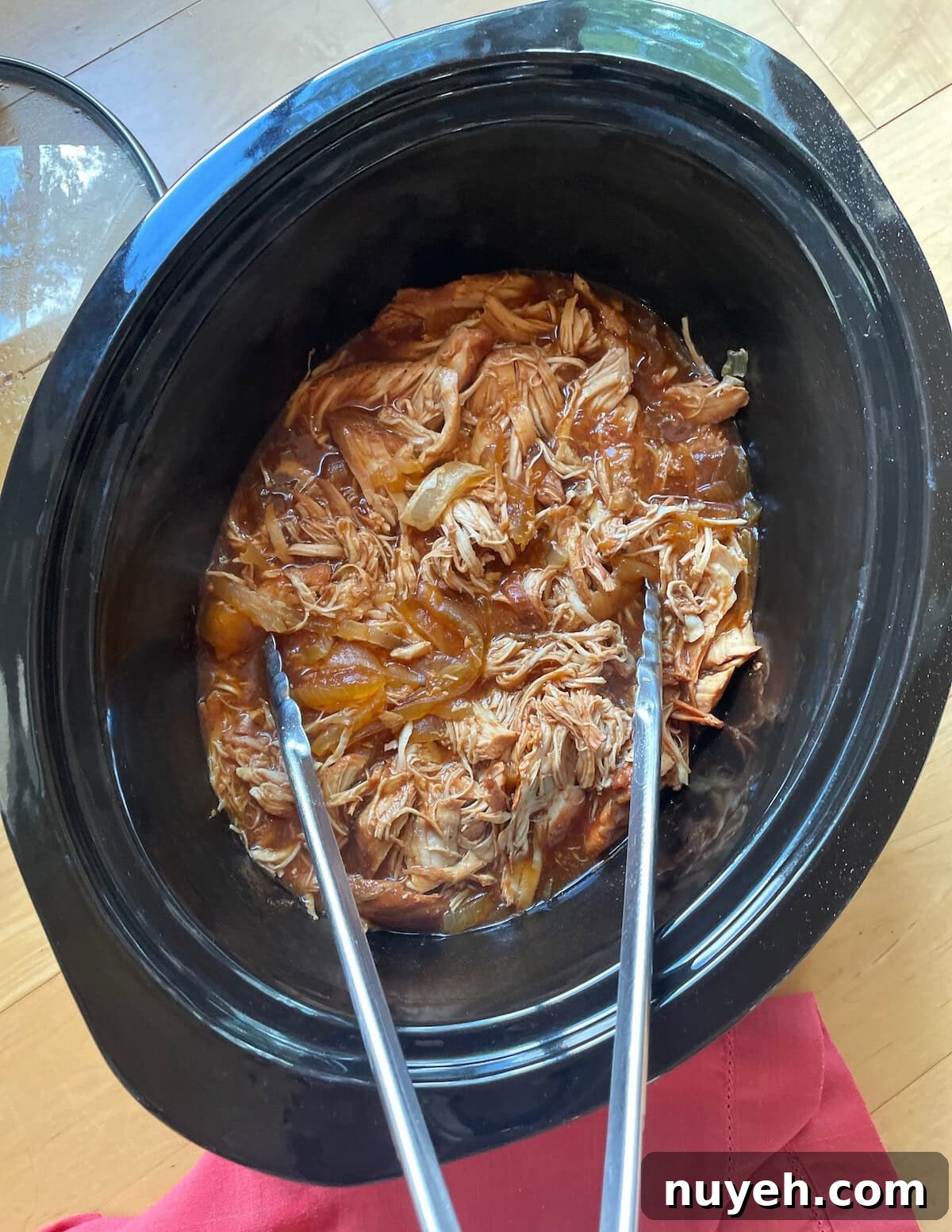 A top-down view of shredded pulled chicken in a black crock pot with tongs, resting on a wooden surface with a red cloth underneath.