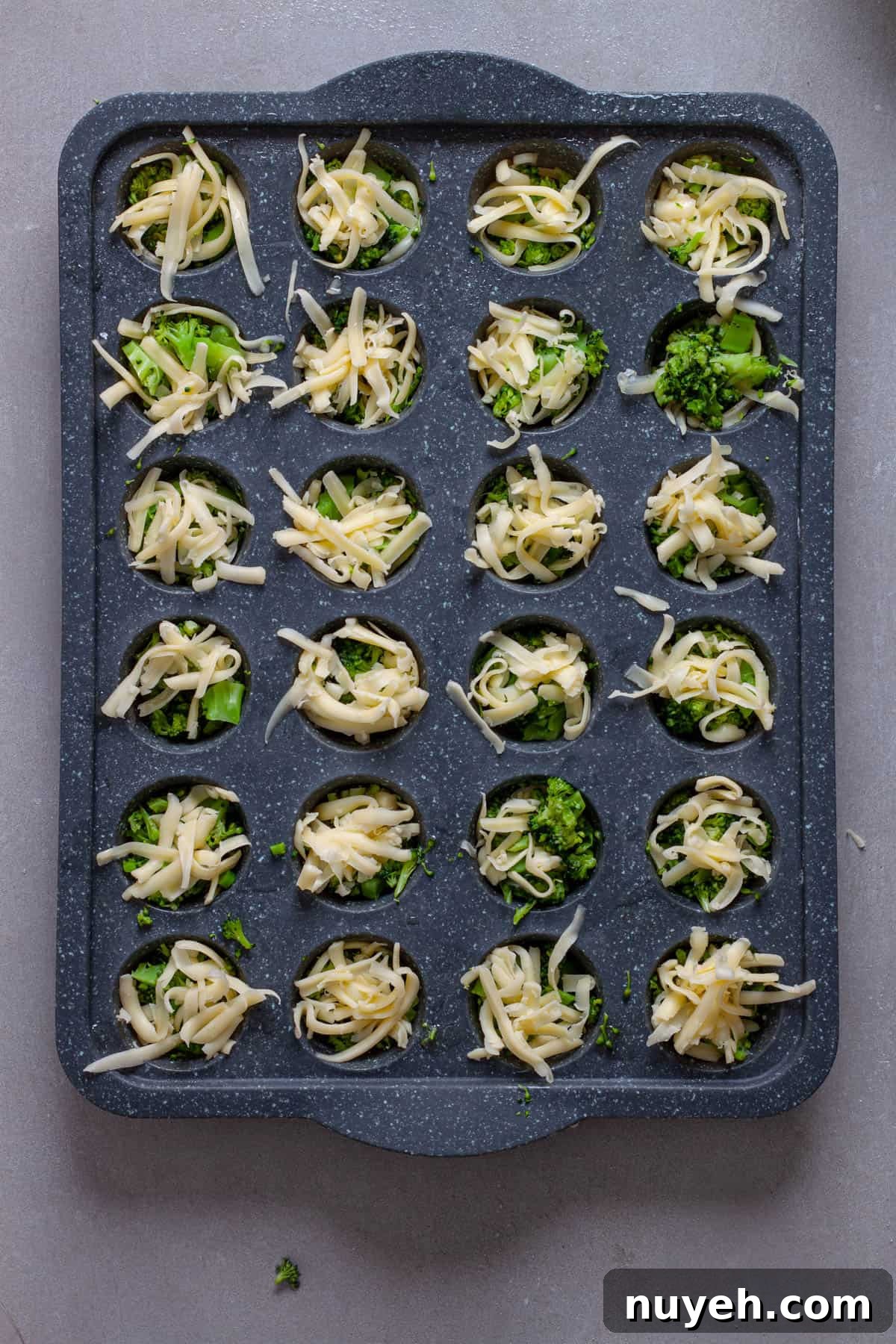 A mini muffin pan with shredded cheese and chopped broccoli.