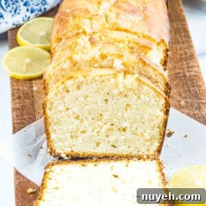 Close-up of a lemon bread loaf with clean slices, showcasing the delicate crumb and shiny glaze, accompanied by lemon halves and a blue-patterned cloth.