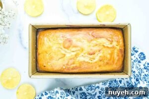 A fully baked lemon bread in a loaf pan, golden brown on top, cooled and ready to slice, with halved lemons beside it for garnish.