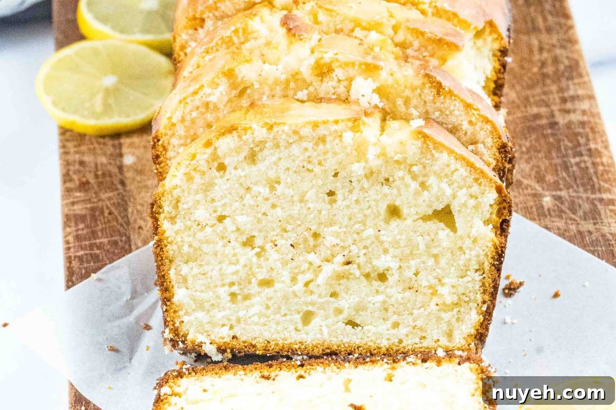Side view of sliced lemon bread on parchment paper, revealing its airy texture and glazed crust, with lemon halves on a rustic wooden board in the background.