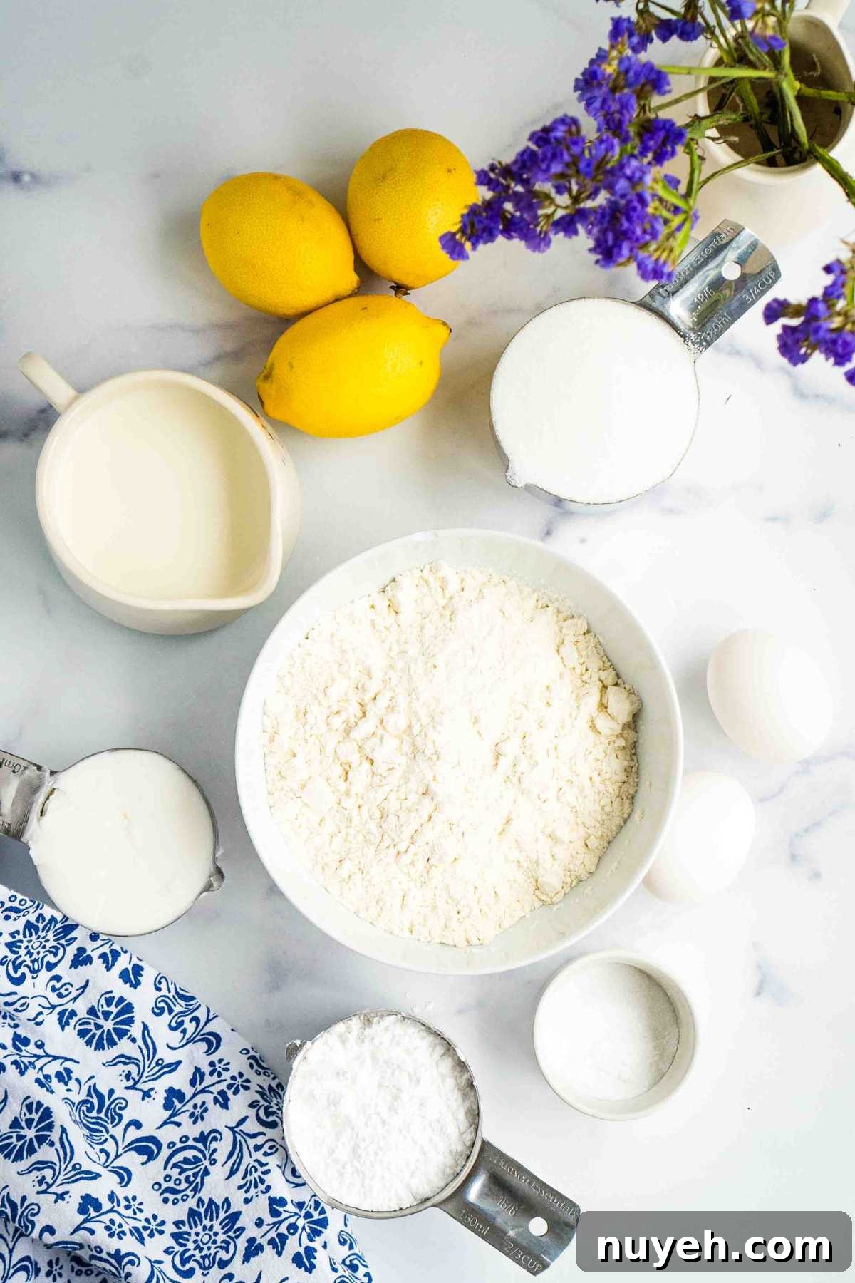 Ingredients for lemon bread neatly arranged on a marble countertop, including lemons, milk, sugar, and flour, ready for preparation, with a touch of purple flowers for decoration.