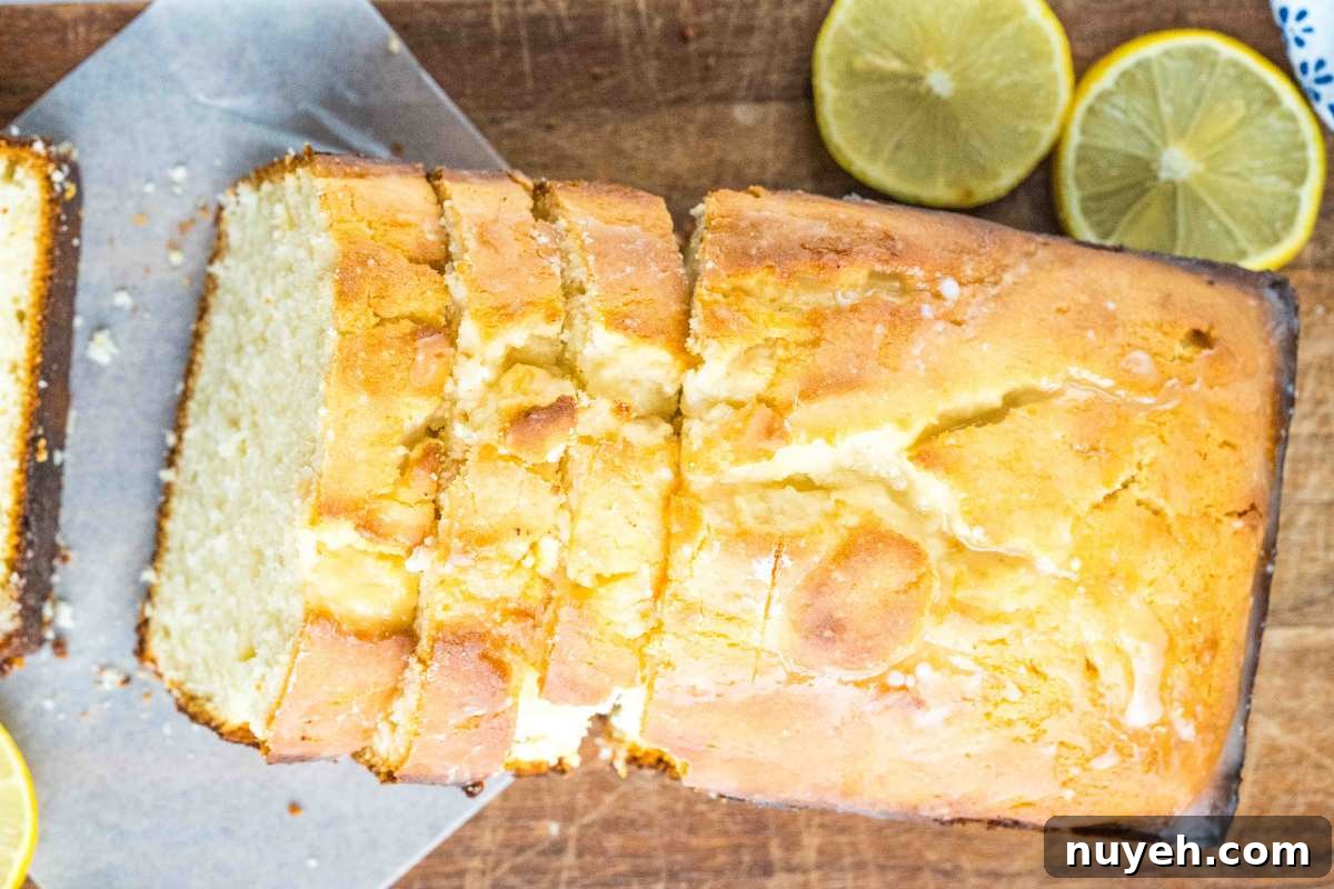 Overhead view of a lemon bread loaf with several slices cut, displaying a moist interior with a golden-brown crust, garnished with lemon halves on a wooden surface.