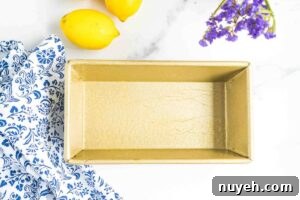 An empty, greased loaf pan next to fresh lemons and a decorative blue and white cloth, set up for pouring in lemon bread batter.