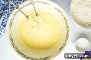An electric mixer in a bowl containing a creamy, pale yellow batter, indicative of a mixture of sugar, shortening, and eggs, with ingredients in the background.