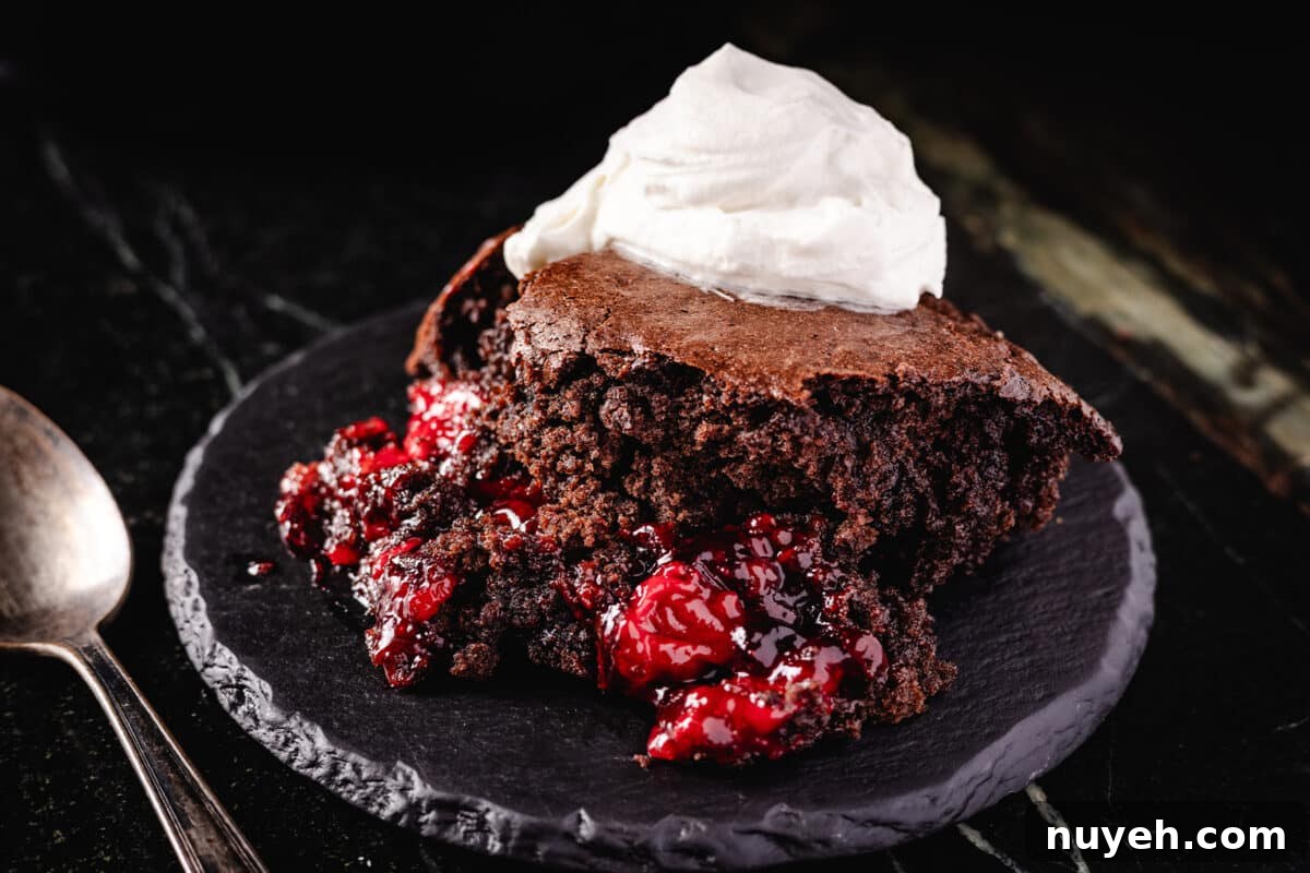 A chocolate berry cobbler topped with whipped cream resting on a black stone platter next to a metal spoon.