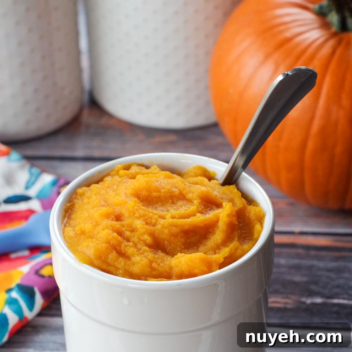 White canister with pumpkin puree and spoon, whole pumpkin in background.