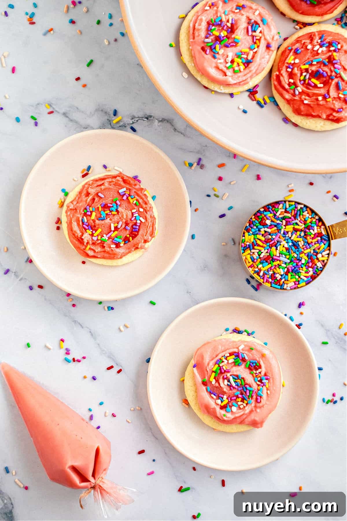 Classic Frosted Sugar Cookies Recipe 11 Top view of white plates with pink-frosted sugar cookies. Cup with sprinkles. Marbled surface.
