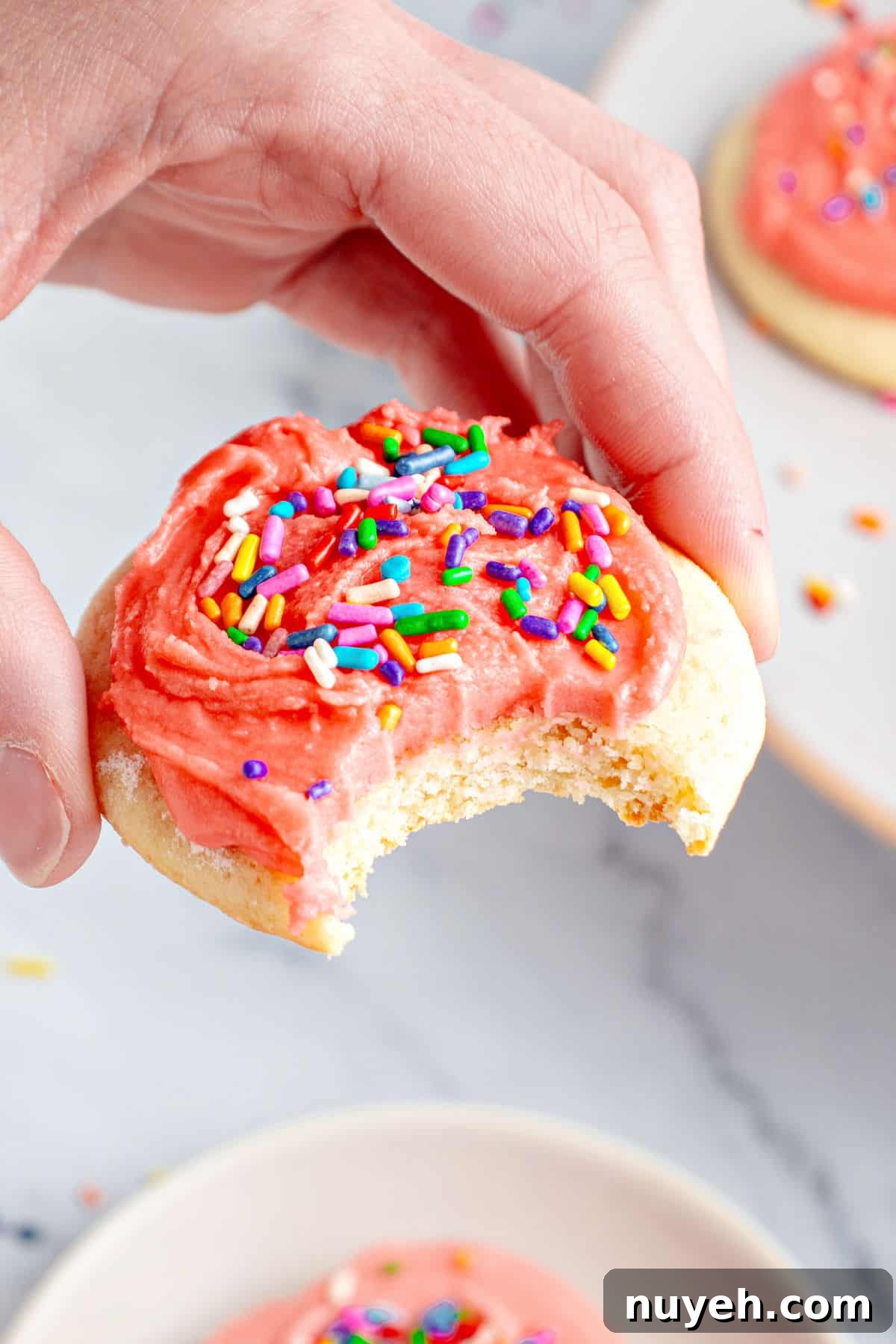 Classic Frosted Sugar Cookies Recipe 3 Hand holding bitten pink-frosted sugar cookie with sprinkles. Marbled surface.