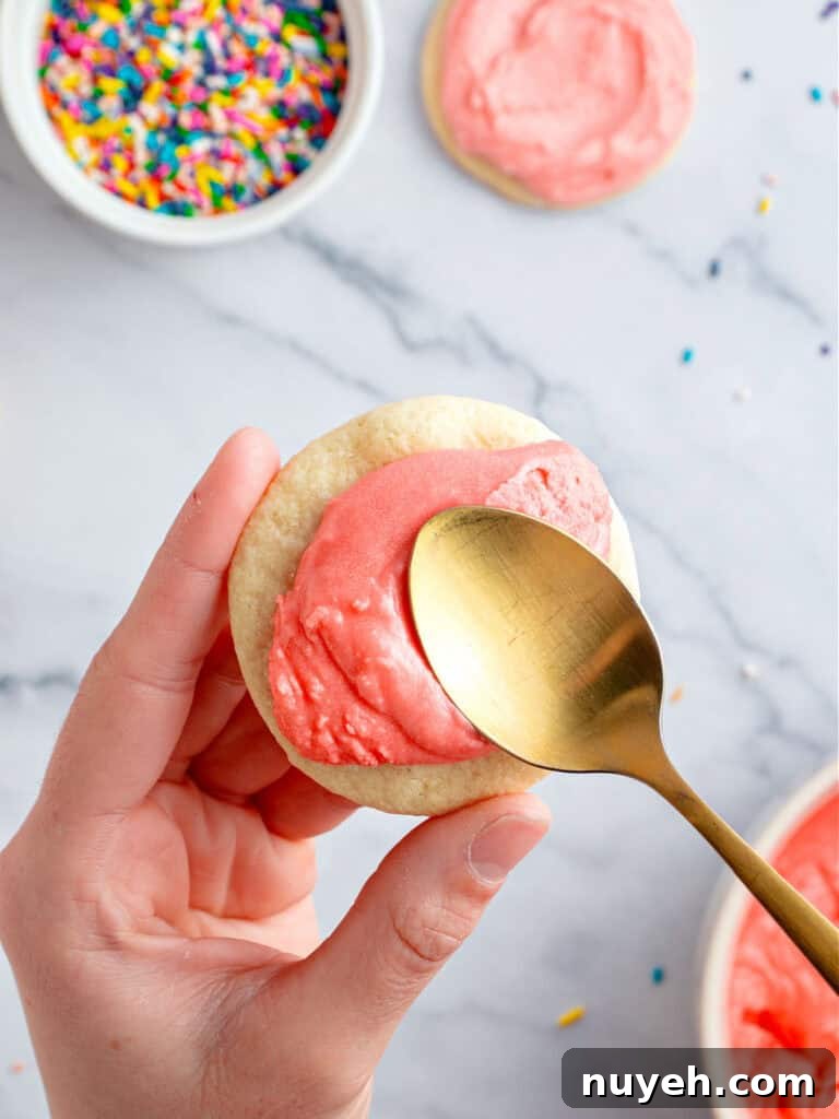 Classic Frosted Sugar Cookies Recipe 9 Spreading pink frosting with a gold spoon on a round sugar cookie. White marbled surface.