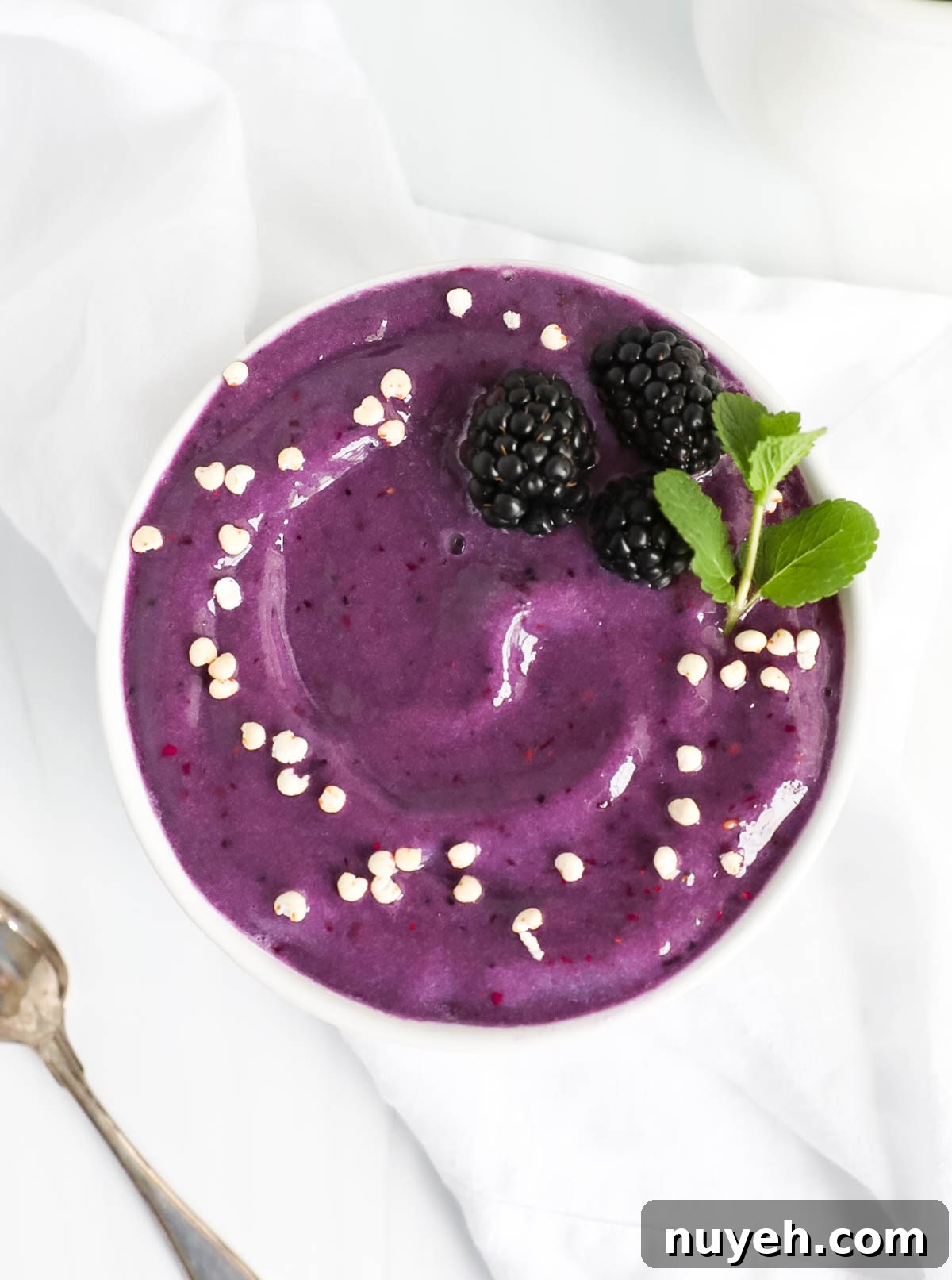 A purple smoothie in a white bowl garnished with three blackberries, fresh mint, and white puffed cereal.