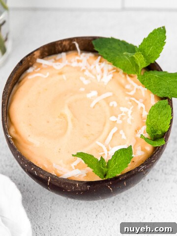 A brown coconut bowl with an orange smoothie inside. The smoothie is garnished with coconut flakes and fresh mint.