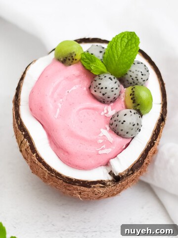 A pink dragon fruit smoothie in a fresh coconut. Garnished with balls of dragon fruit, kiwi, and fresh mint.