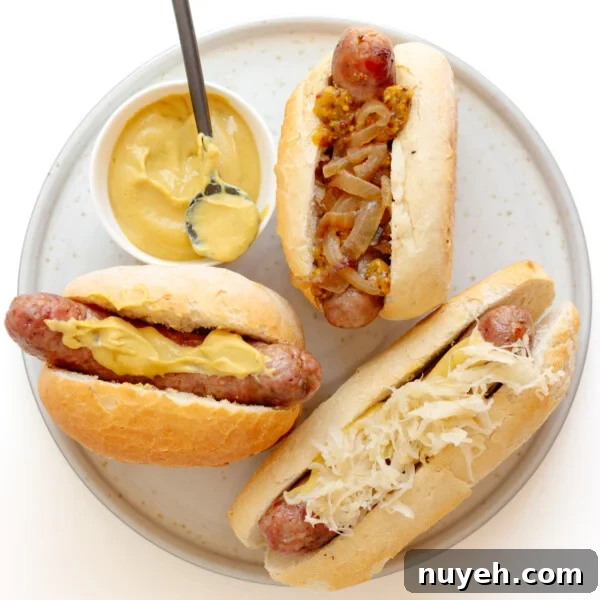 Three grilled bratwurst on buns with different toppings sitting on a plate with a small bowl of mustard.