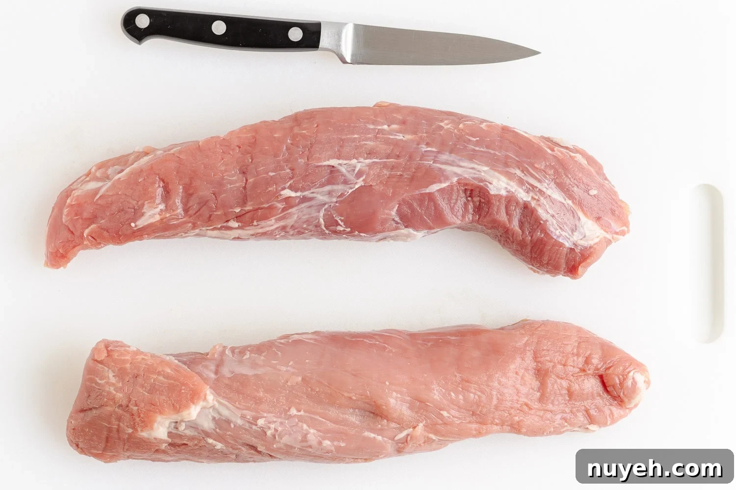 Two trimmed pork tenderloins on a white plastic cutting board with small knife.
