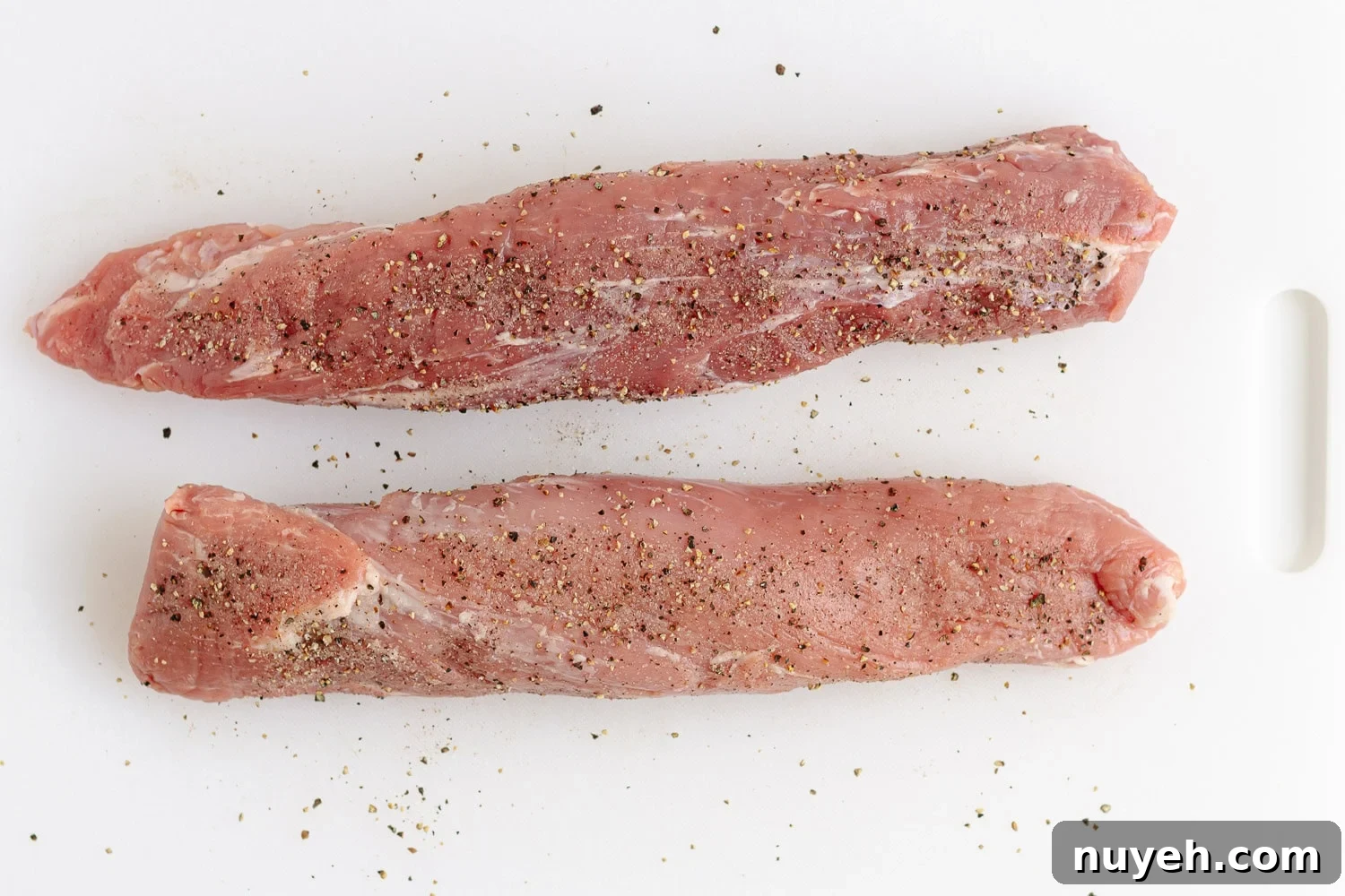 Two raw pork tenderloins on a white plastic cutting board seasoned with salt and pepper.