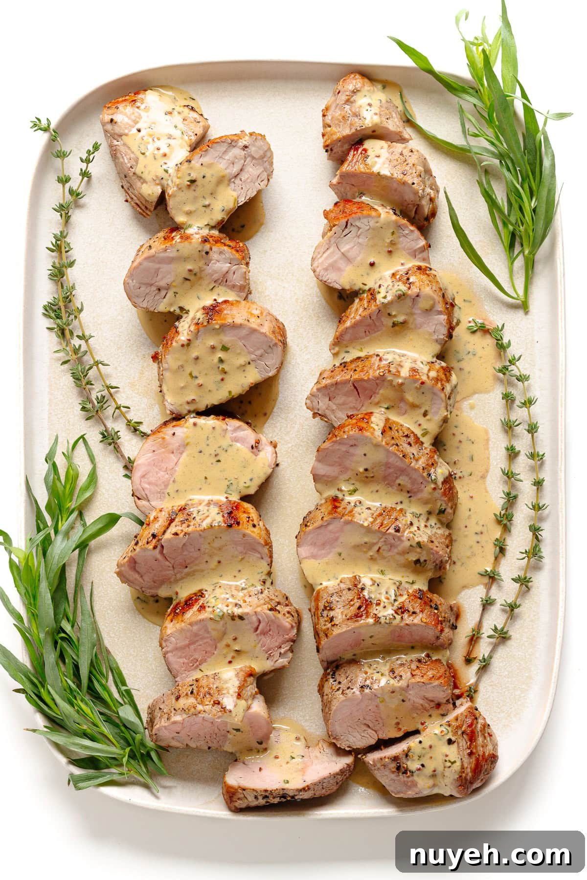 Platter of sliced pork tenderloin drizzled with creamy mustard sauce and garnished with fresh sprigs of thyme and tarragon.