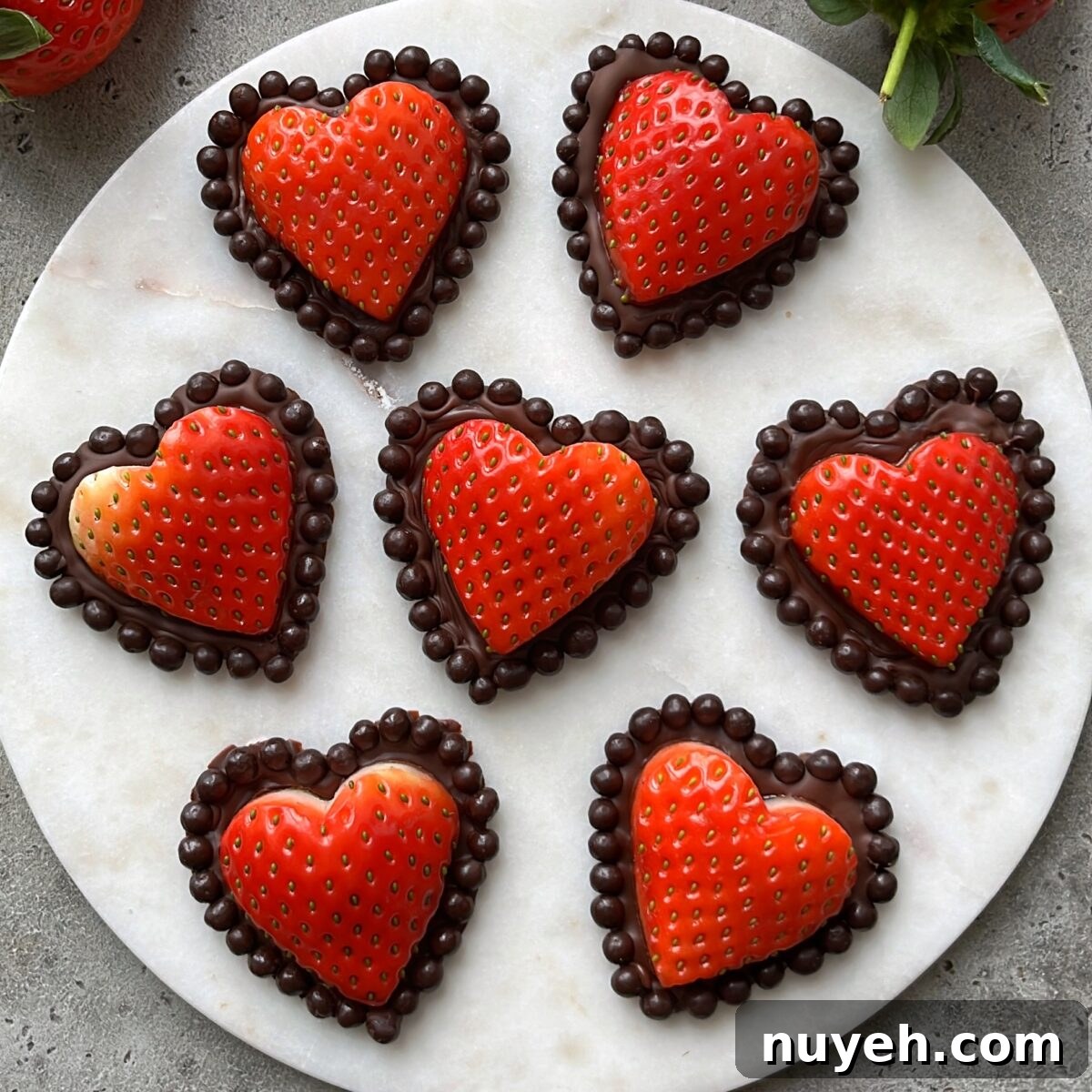 Seven heart-shaped strawberries dipped in chocolate and bordered with chocolate balls are arranged on a round white marble surface.