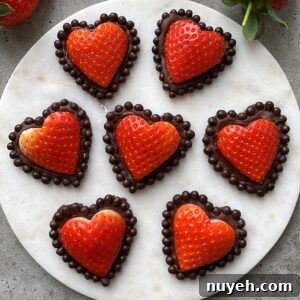 Seven heart-shaped strawberries dipped in chocolate and bordered with chocolate balls are arranged on a round white marble surface.