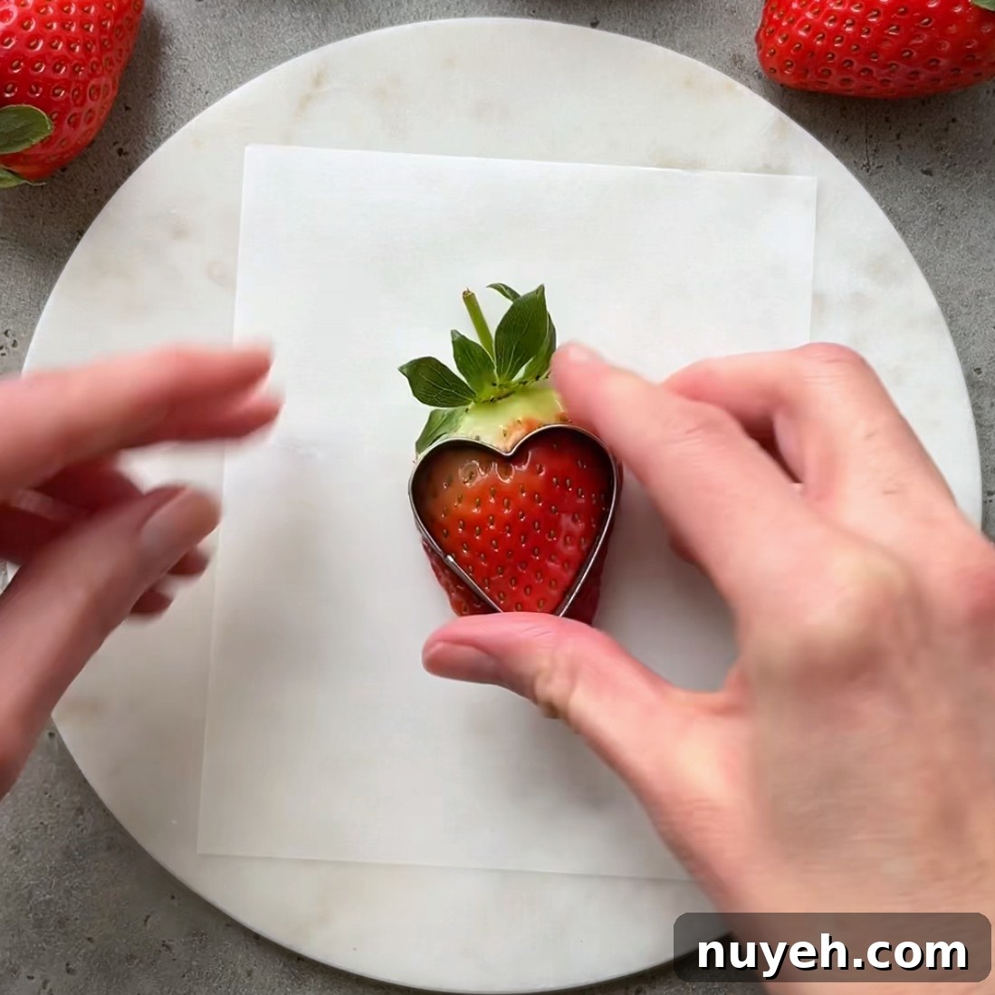 A hand uses a cookie cutter to cut out a heart from of a strawberry half.