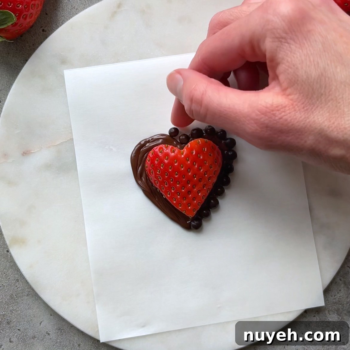 A hand places chocolate ball sprinkles in the dark chocolate outline surrounding a strawberry heart.