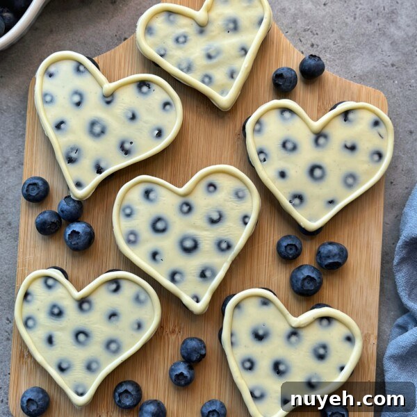 Six heart-shaped cheesecakes with embedded blueberries are arranged on a wooden board, creating delicious bites surrounded by fresh blueberries on a gray surface. Perfect for anyone who loves health-conscious food recipes.