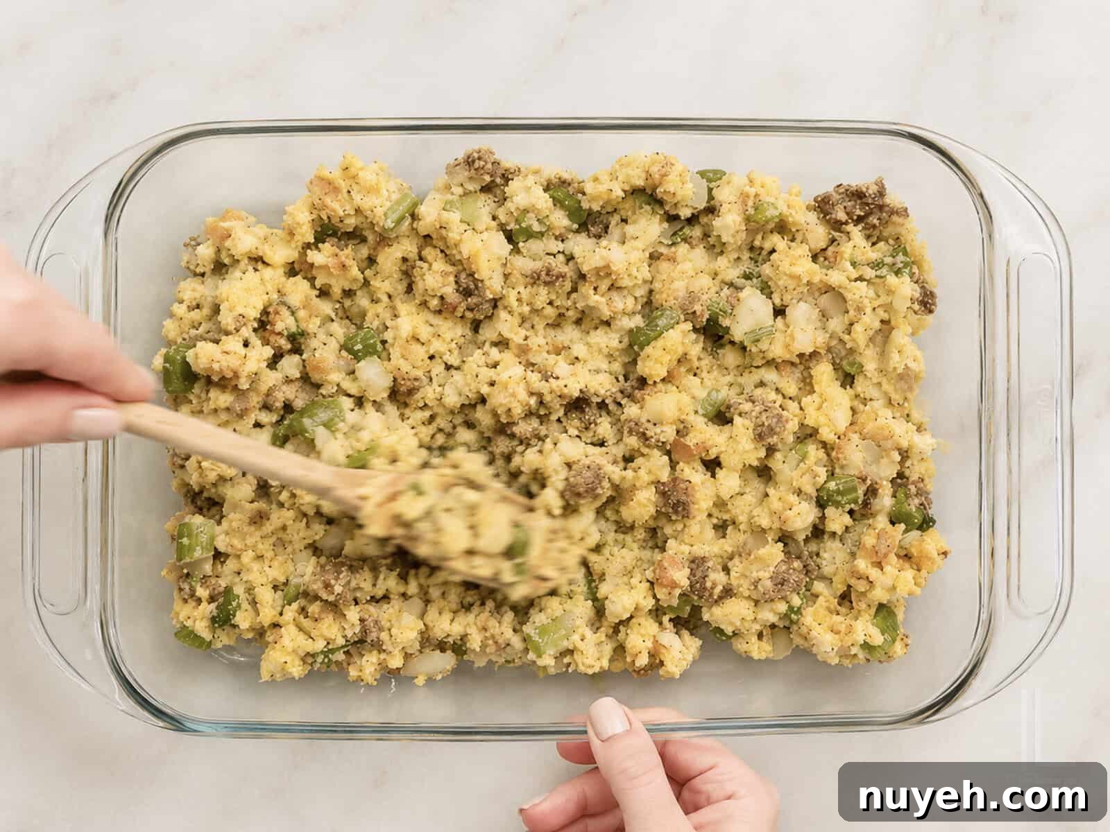 Unbaked Jiffy cornbread dressing with sausage in a glass dish, being spread for baking.