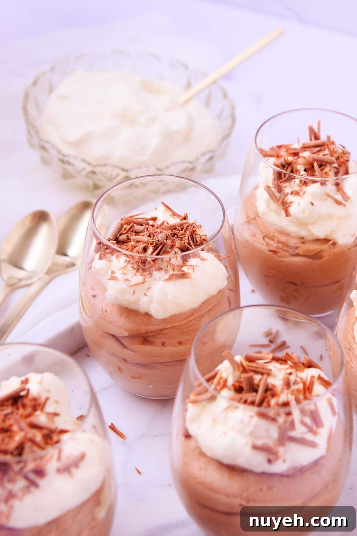 Four glasses of milk chocolate mousse, with a small glass bowl of whipped cream, and gold teaspoons in the background.